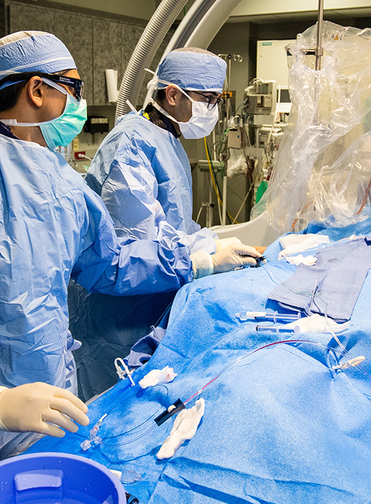 Two neurointerventional radiologists operate on a patient under a blue sheet.