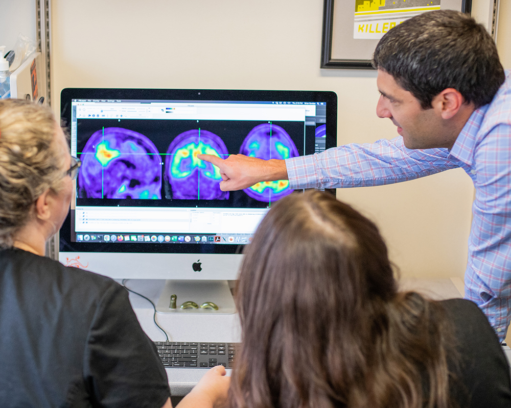 Brian Gordon, PhD, points to a colorful brain scan on a computer screen with two onlooking researchers in the Neuroimaging Labs Research Center.