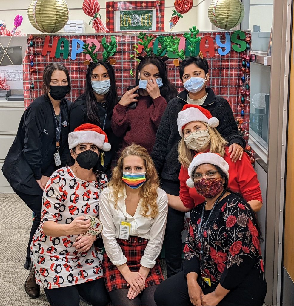 Eight members of the Benzinger lab pose in two rows. All are wearing masks, and some are wearing green reindeer antlers, while others wear red and white Santa Claus hats. The lab members in the front row are dressed in red and green holiday outfits.