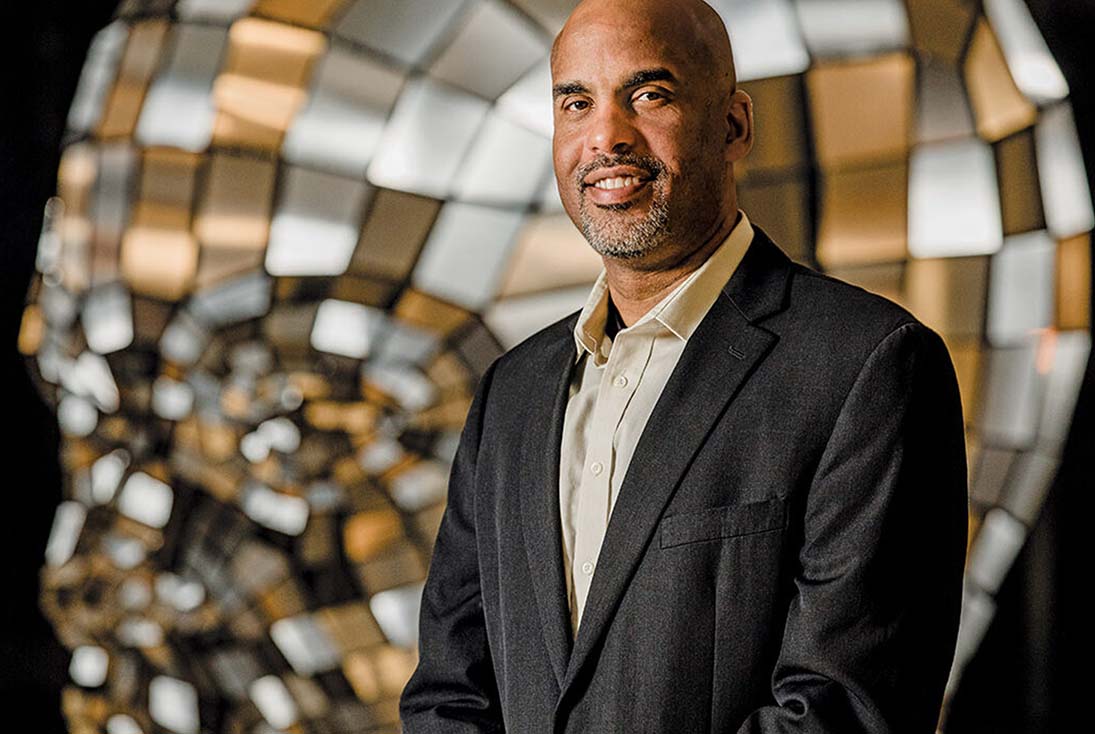 Neuroscience alumnus Damien Fair, PhD, stands in a gray suit infront of a silver and bronze mosaic.