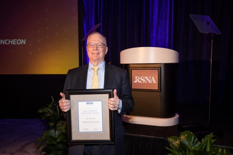 Professor Emeritus Dennis Balfe, MD, stands in front of a podium with his Outstanding Educator Award plaque at the 108th RSNA Annual Meeting in Chicago.