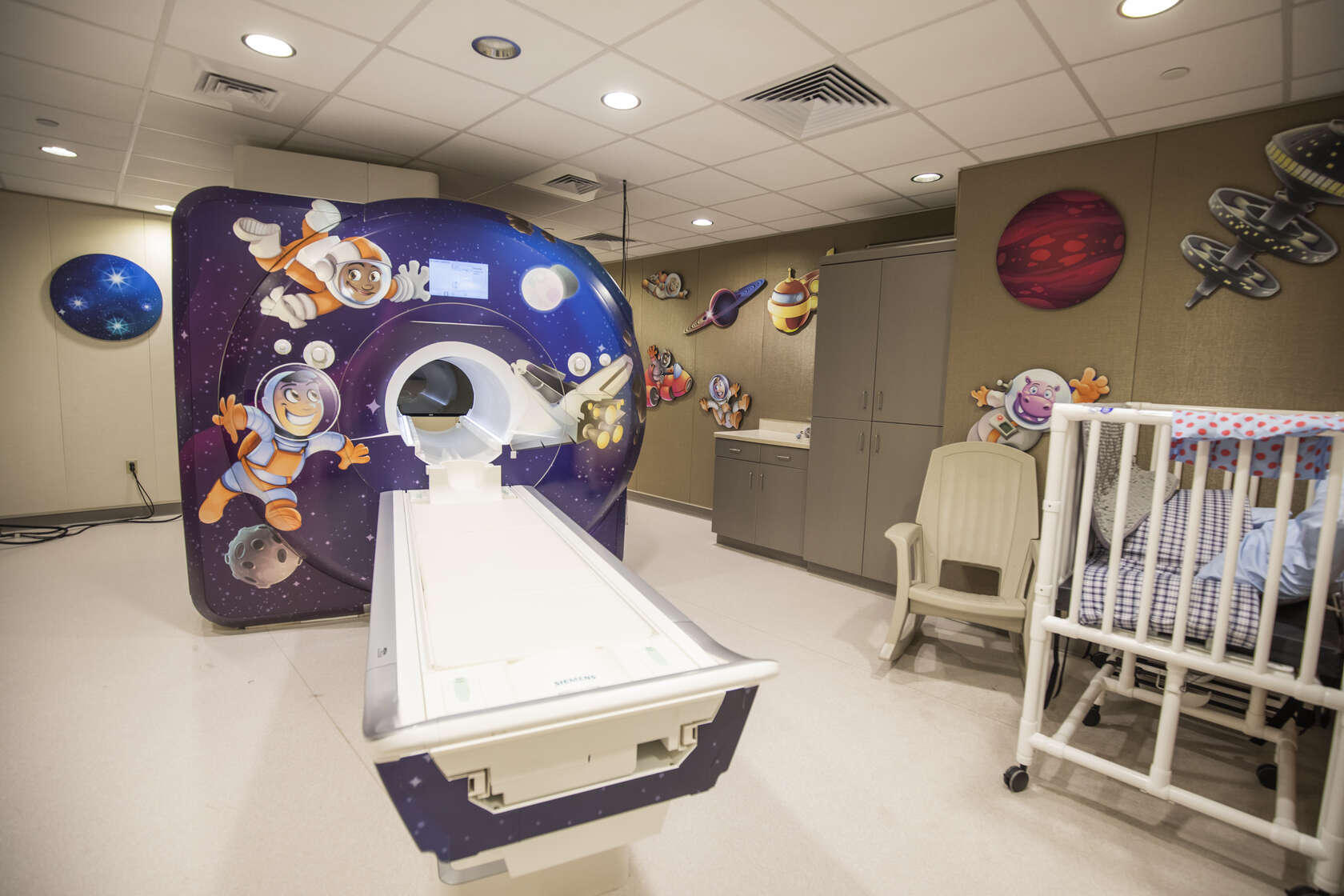 A 3T MRI scanner in an imaging exam room is decorated with a space-themed design on the scanner and decals around the room.
