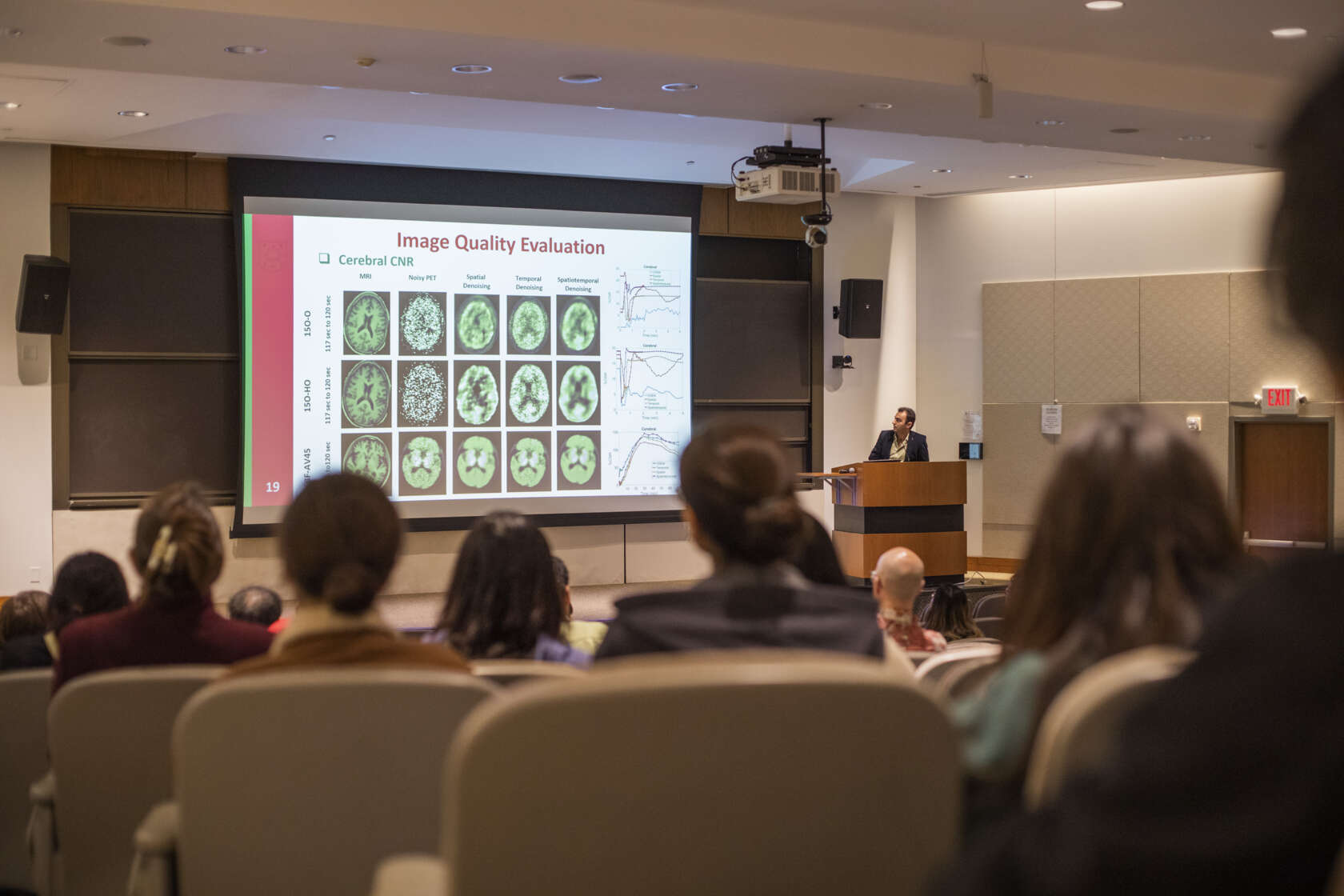 A group of people sitting in Moore Auditorium look at a speaker at a podium and a projector screen depicting colorful brain scans at the Imaging Sciences Pathway Retreat on March 31, 2023, at Washington University School of Medicine in St. Louis.