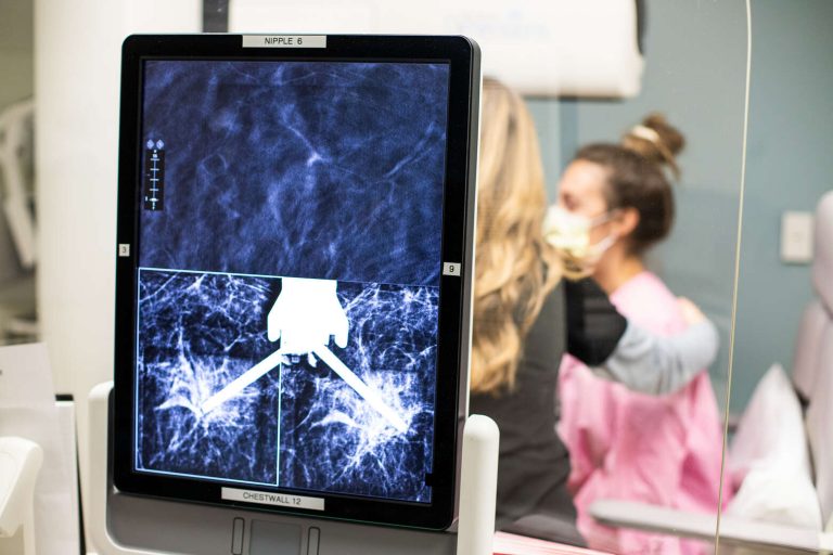 A screen depicting a breast imaging scan in the foreground while a patient and a nurse interact in the background.