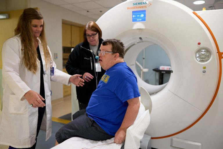 A down syndrome patient sits on the bed of a CT scanner while a technician helps him get ready for a scan