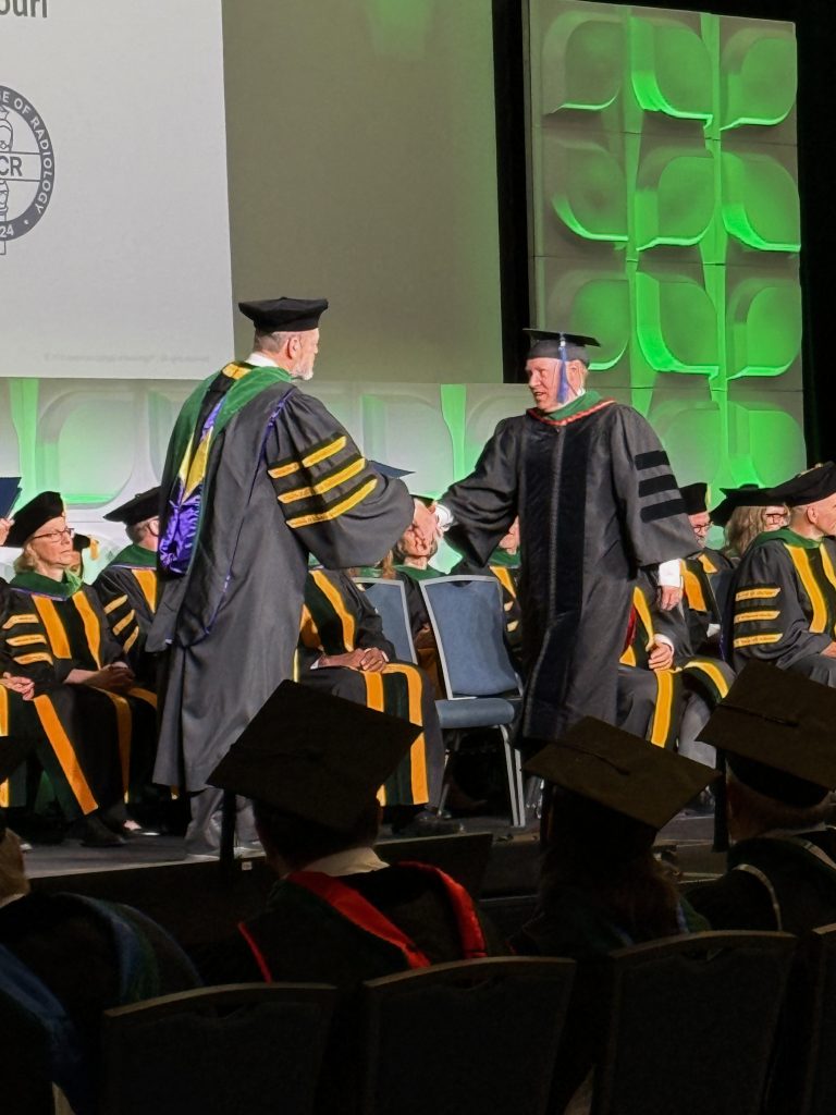 Two men in graduation caps and gowns shake hands on stage during the ACR convocation ceremony.