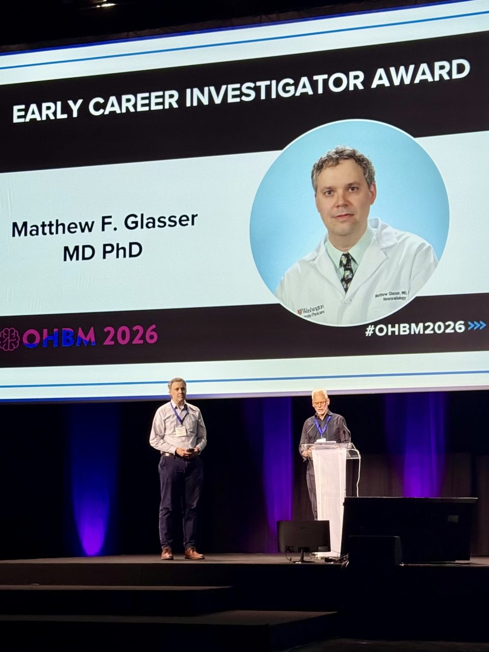 An awards presentation at the 2026 OHBM Annual Meeting. A large screen displays "Early Career Investigator Award" along with a photo and text: "Matthew F. Glasser MD PhD." Two individuals stand on stage beneath the screen.