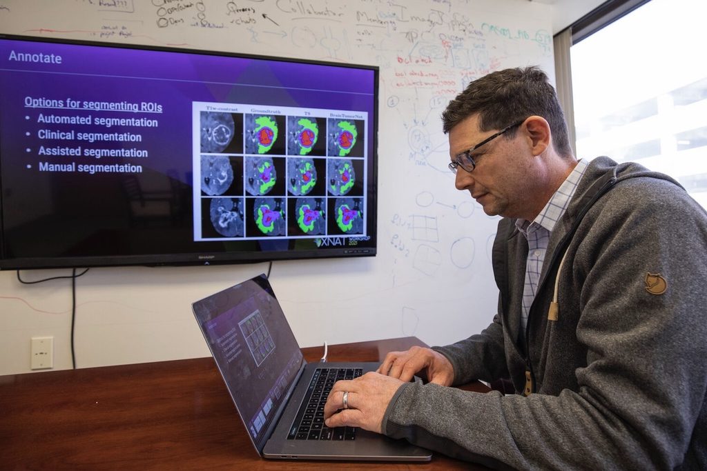 Daniel Marcus, PhD, is working on a laptop in an office. There is a screen on the wall displaying medical imaging titled "Options for segmenting ROIs," with labeled categories: Automated, Clinical, Assisted, and Manual segmentation. The wall has various notes and diagrams.